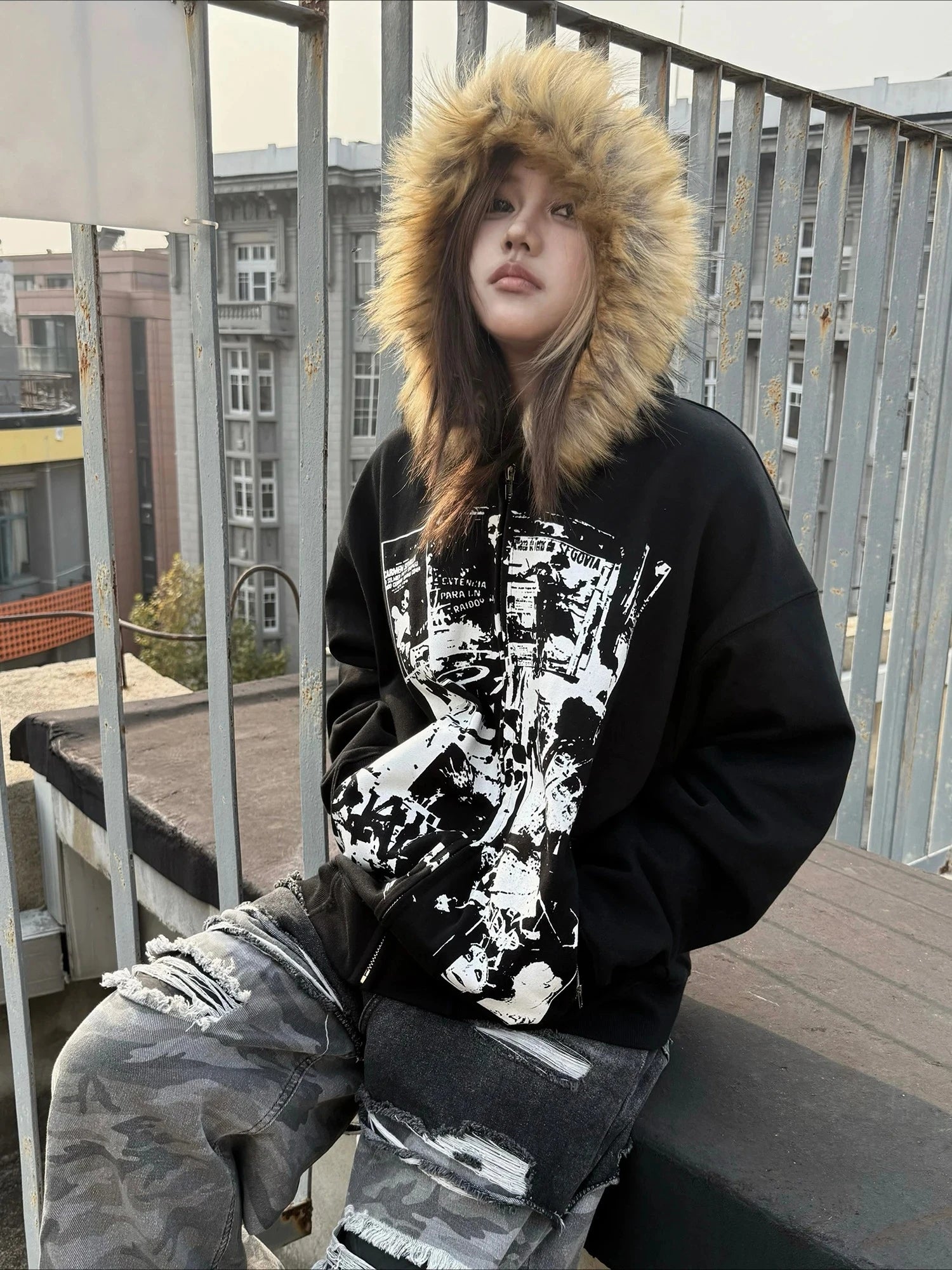 Person wearing a black hoodie with a graphic design and fur-lined hood on a balcony.